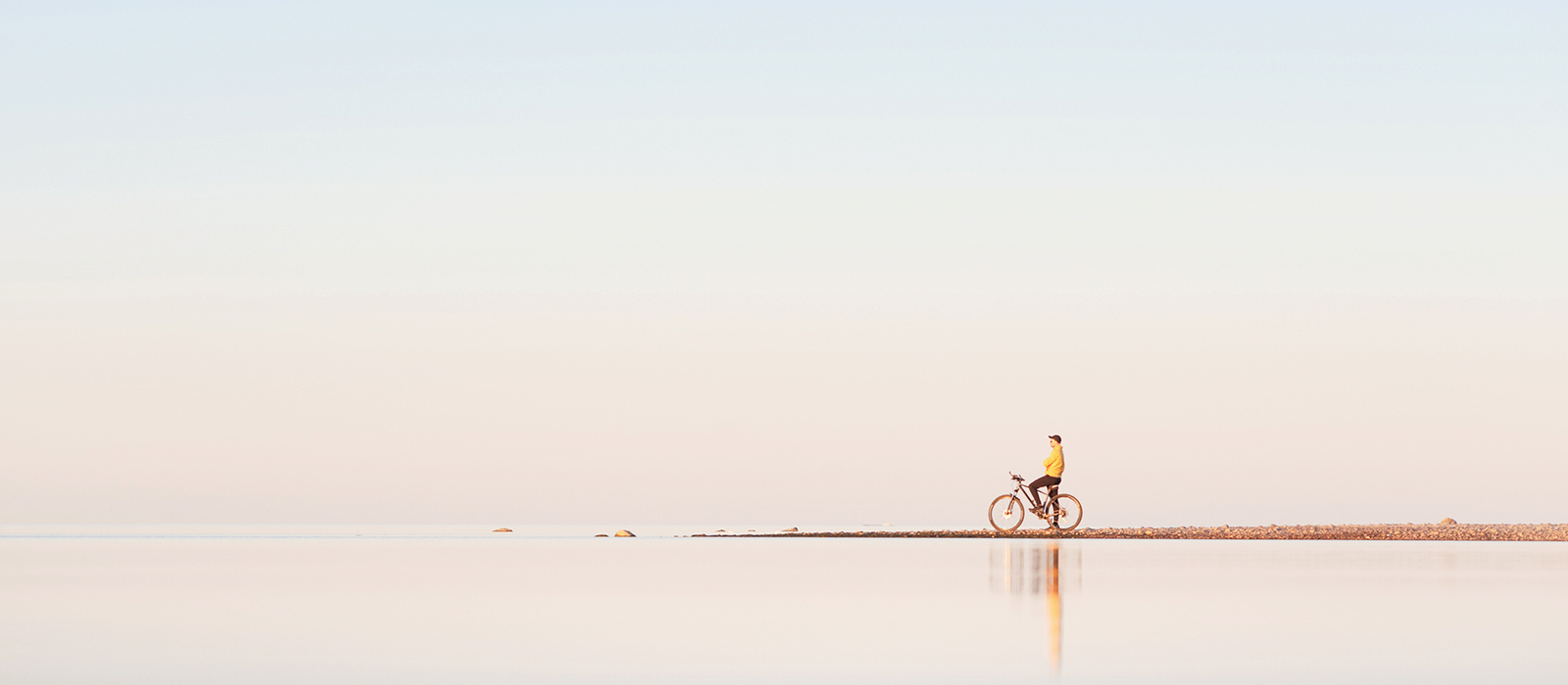 Decorative. A lone cyclist stands still on a bicycle on a narrow strip of land surrounded by a calm reflective water surface.